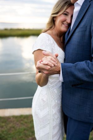 A romantic close-up of a couple holding hands, with a white lace dress and navy suit against a waterfront backdrop.