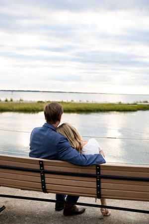 A couple sits together on a wooden bench overlooking a serene lake on a cloudy day.