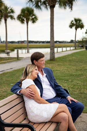 A couple sits together on a wooden bench along a palm tree-lined waterfront path on a sunny afternoon.