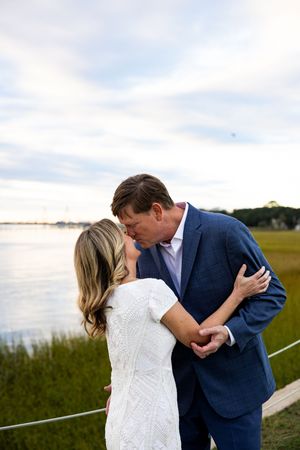 A romantic wedding couple shares an intimate moment by the water during sunset, dressed in white and navy blue.