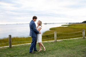 A romantic moment captured between a couple on grassy waterfront with wooden fence and cloudy sky.