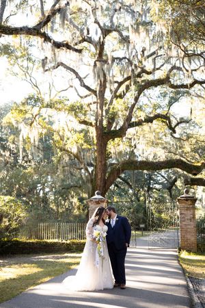 A couple shares an intimate moment under Spanish moss draped oak trees along a sunlit garden pathway.