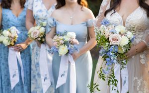 Wedding party in light blue bridesmaid dresses holding white and blush floral bouquets stand together outdoors.