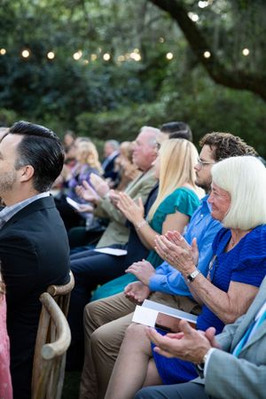 Wedding guests seated outdoors clap and celebrate during an evening ceremony with string lights overhead.