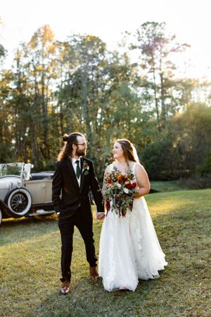 A wedding couple stands on grass during golden hour with a vintage car in the background.