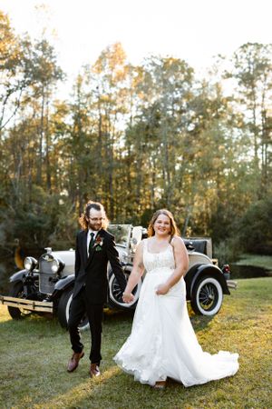 A joyful wedding couple poses next to a vintage car on their special day in an outdoor autumn setting.