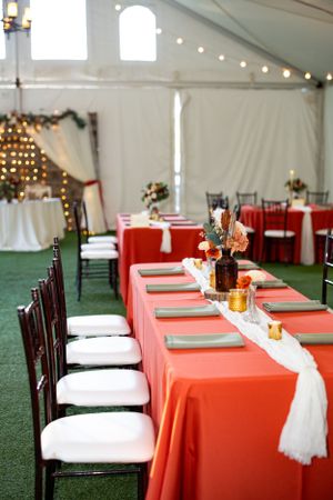 Wedding reception setup with coral tablecloths, white chairs and string lights in an elegant tent venue.
