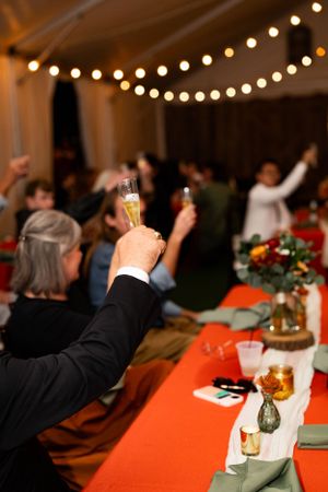 A hand raises a champagne glass during a festive celebration with string lights and red tablecloths.