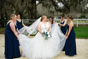 Bridesmaids in navy dresses help arrange a bride's flowing veil in an outdoor setting.