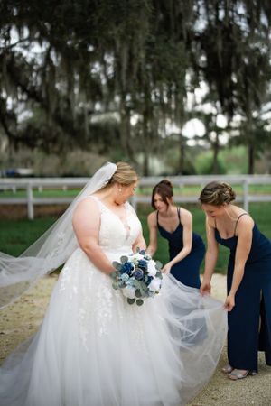 Bridesmaids in navy dresses assist with the bride's tulle gown outdoors.