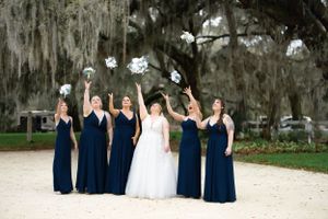Wedding party in navy blue dresses and white gowns celebrates outdoors under moss-draped trees.