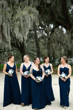 Five bridesmaids in elegant navy blue dresses share a laugh while holding blue and white bouquets.