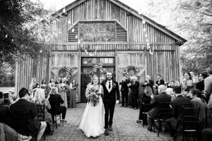 A black and white photograph of a rustic barn wedding ceremony with guests seated outdoors as the couple exits the venue.