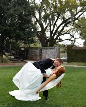 A couple in wedding attire performs a romantic dip pose on a grassy lawn with trees in the background.