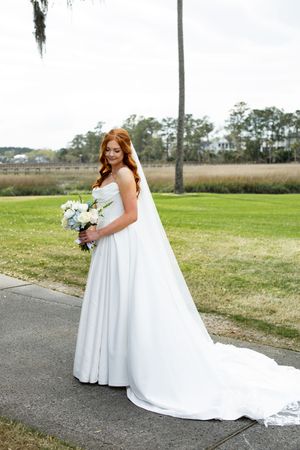 A bride in a flowing white wedding dress walks along a path with a bouquet near a grassy marsh landscape.