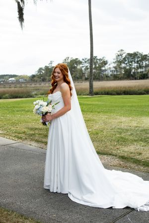 A bride in a flowing white wedding gown stands outdoors on a path with grassy fields and palm trees in the background.