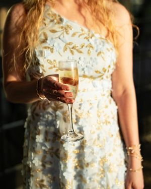 A person wearing a cream-colored dress with floral details holds a glass of wine in a dimly lit setting.