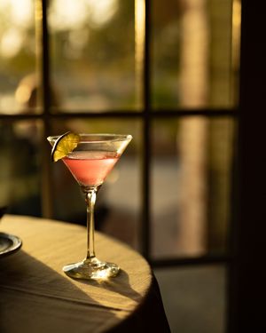 Pink cocktail in martini glass with lemon garnish illuminated by warm sunset light through window.