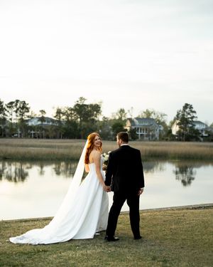 A bride and groom walk along a serene lakeside path at sunset with their backs to the camera.