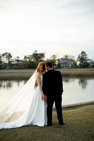 A romantic lakeside wedding scene with a couple in formal attire embracing at sunset.