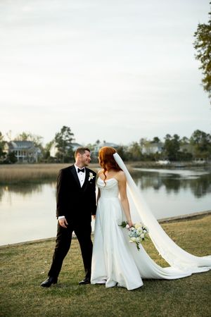 A bride and groom stand together on a grassy lawn beside a tranquil lake at sunset.