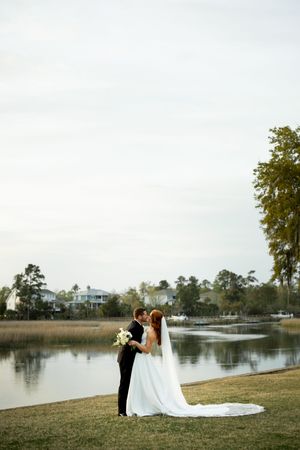 A couple shares an intimate moment by a serene lake with marsh grass and coastal homes in the background.
