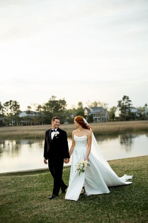 A couple in wedding attire walks along a grassy waterfront during golden hour as their dress flows in the breeze.