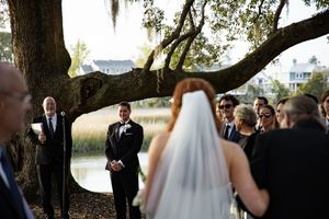 A wedding ceremony takes place under a large oak tree draped with Spanish moss beside a lake at sunset.