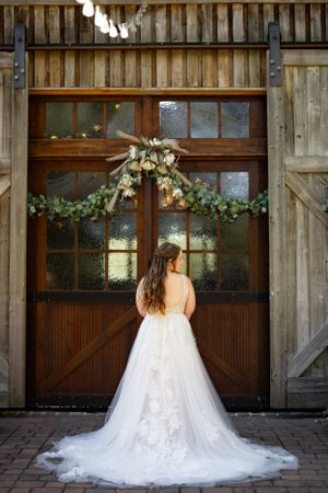 A white wedding gown with a flowing train stands before a rustic wooden barn door decorated with floral arrangements.