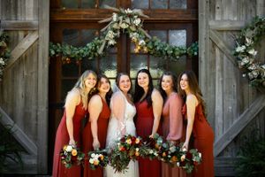 A wedding party wearing burgundy and blush gowns poses in front of a rustic wooden door adorned with greenery.
