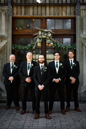 Groomsmen in black suits pose in front of a rustic barn door decorated with floral arrangements.