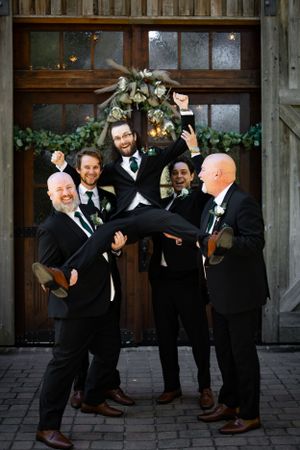 Groomsmen in black suits playfully lift someone in a fun group photo in front of a rustic doorway.