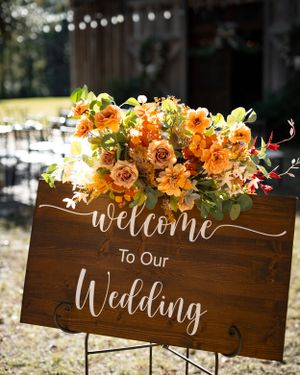 Wooden wedding welcome sign decorated with orange and peach floral arrangements against a rustic outdoor backdrop.