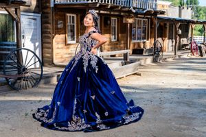 Person wearing an elegant royal blue and silver ballgown poses in front of rustic western buildings.