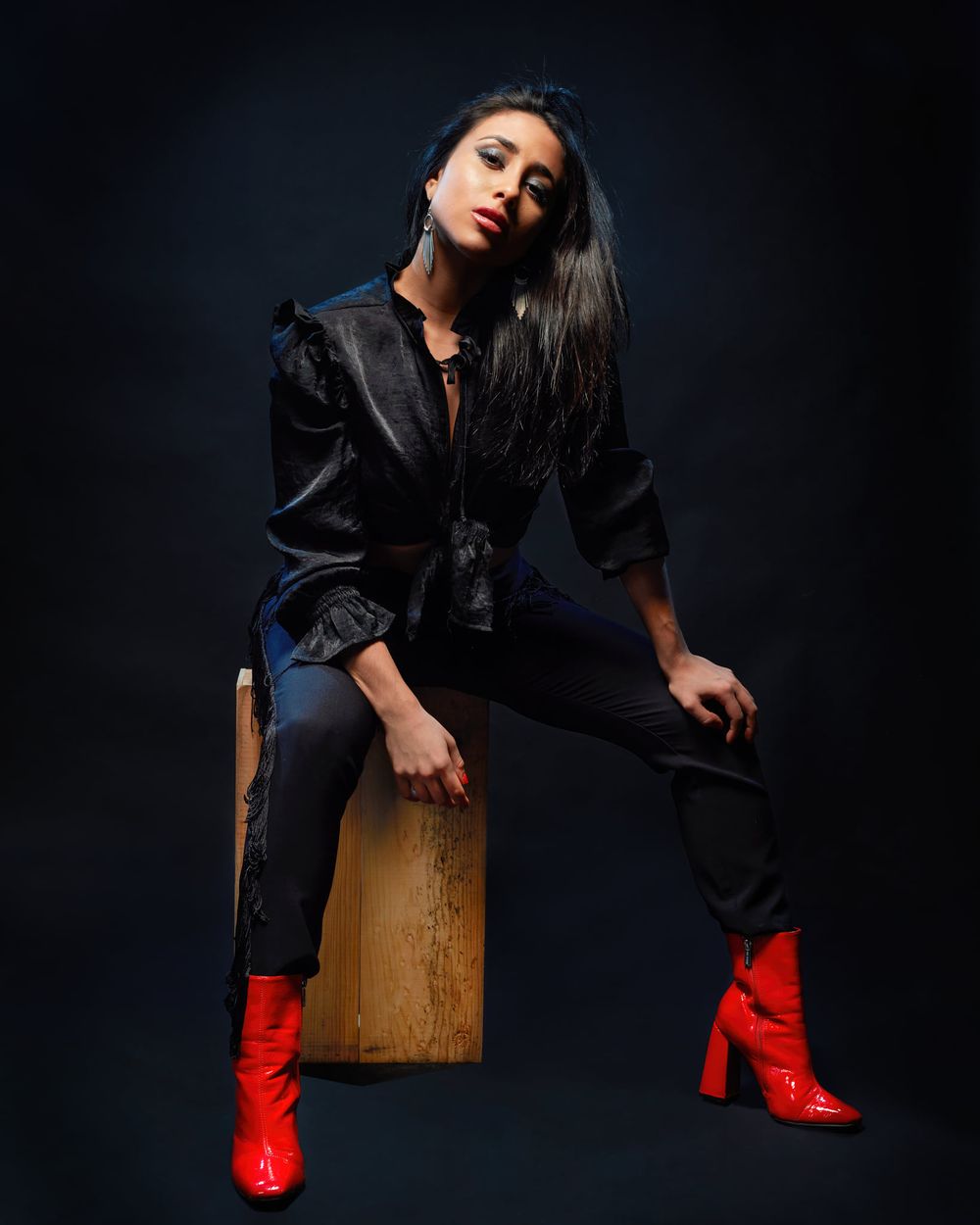 Fashion model poses on wooden block wearing leather jacket and bright red boots against dark background.