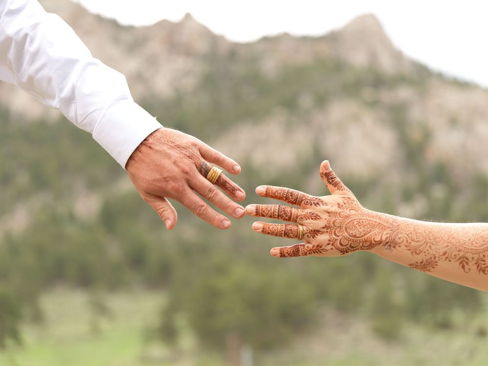 Two hands reaching toward each other, one with a gold wedding ring and one adorned with intricate henna, with mountains in the background.