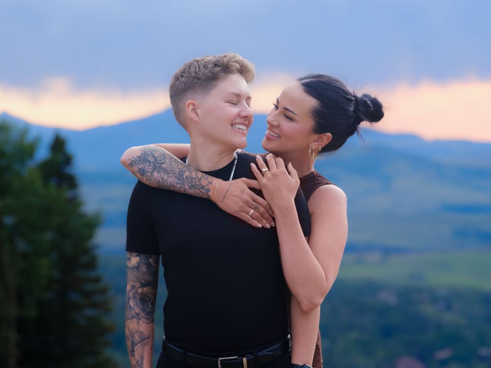 A couple embraces romantically at sunset with mountains and green valleys in the background.