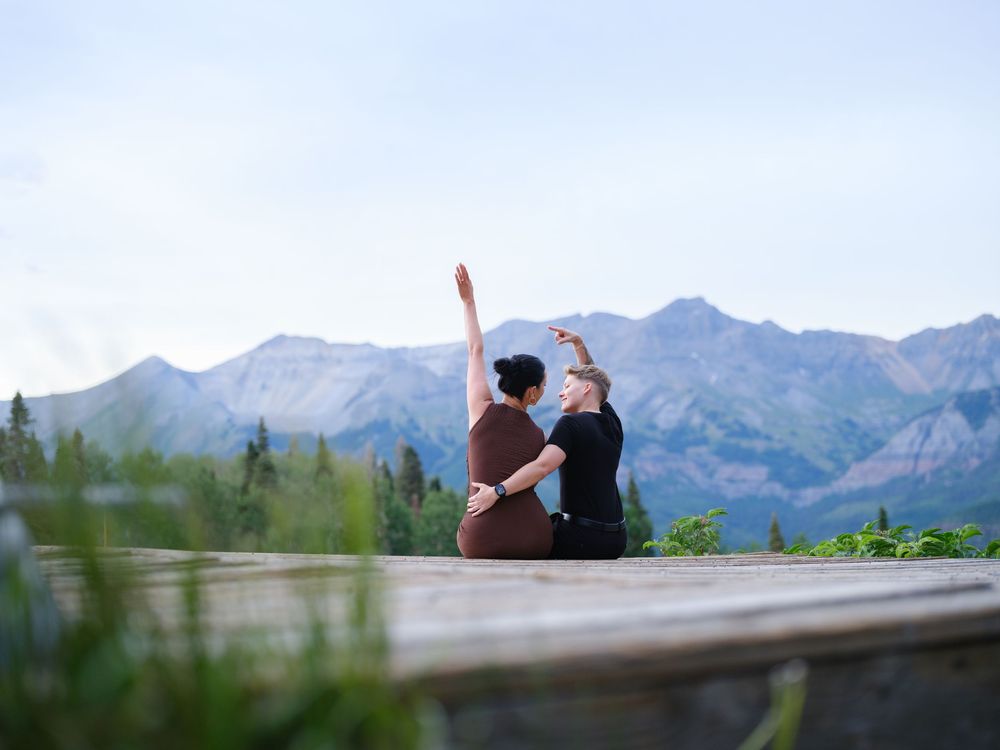 Two people sit together on a wooden deck overlooking majestic mountain peaks in a scenic outdoor setting.