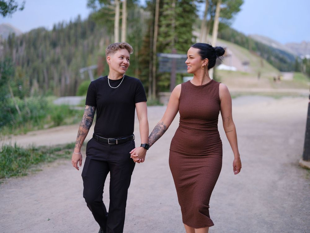 A couple holding hands while walking on a mountain path, one wearing black and the other in a brown dress.