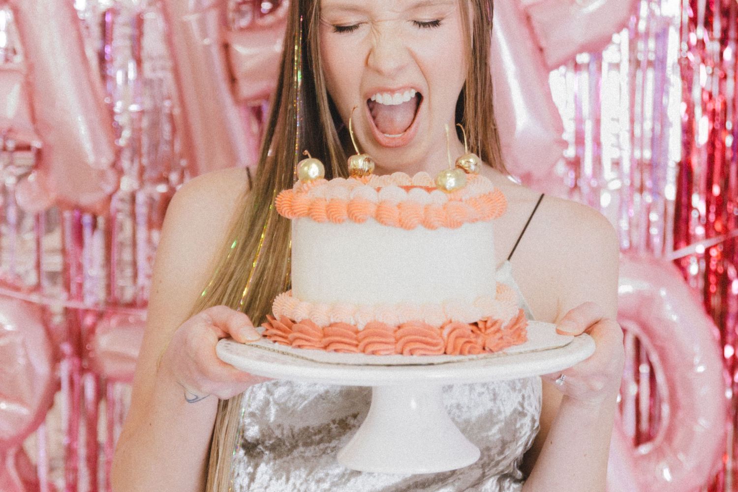Someone holds a white and peach-colored birthday cake with excitement against a pink sparkly backdrop.