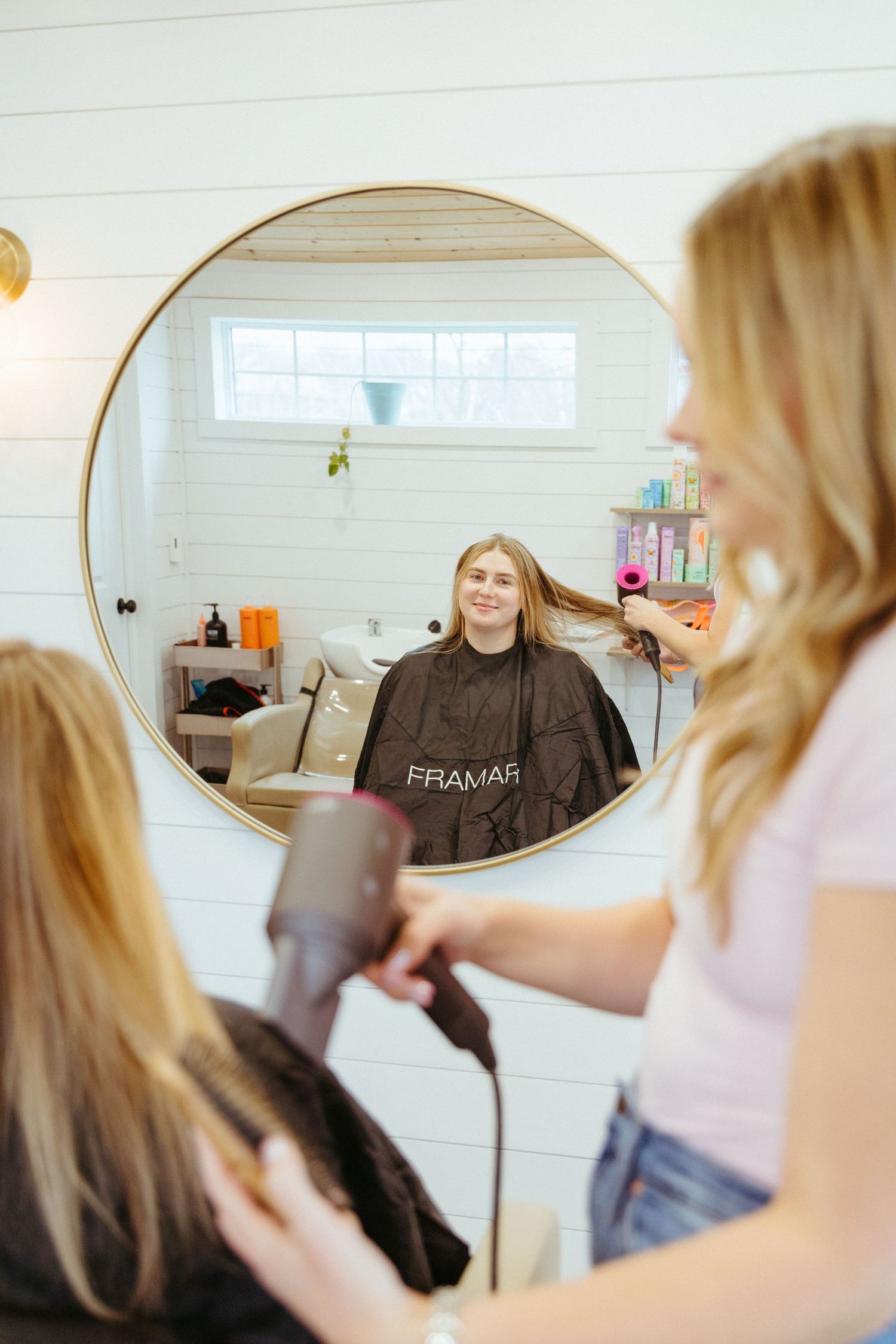 A stylist blow dries a client's hair in a bright salon with a round mirror and white shiplap walls.