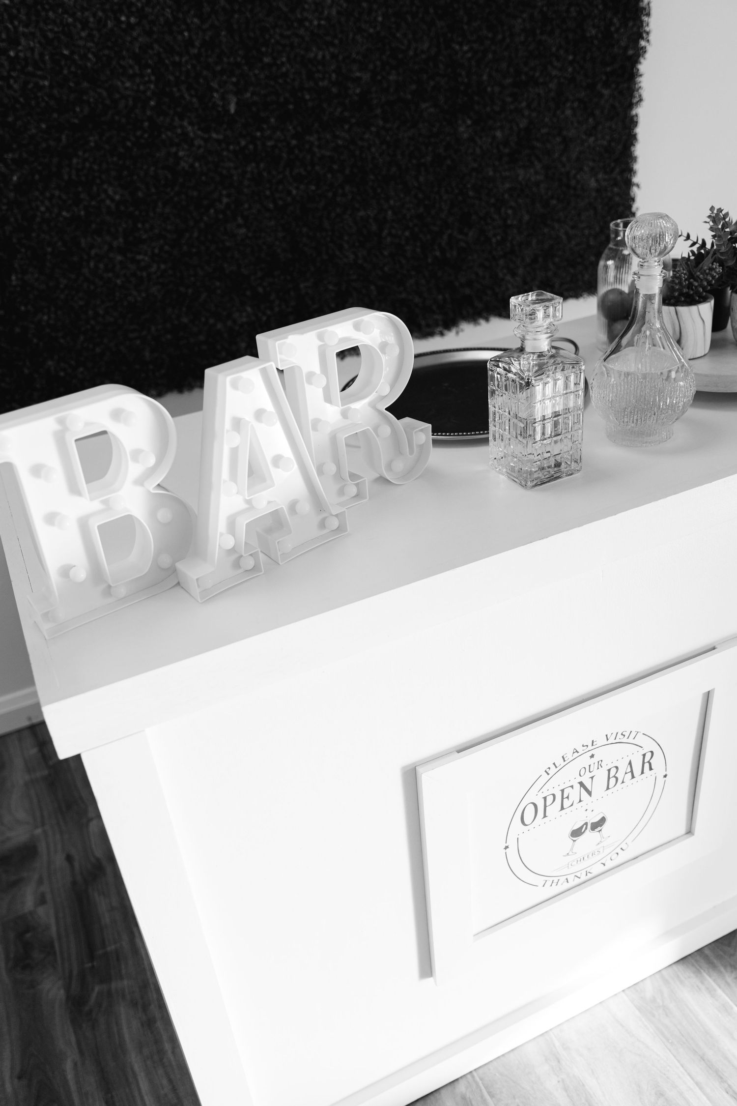 White illuminated BAR letters on top of a white counter with liquor bottles and an Open Bar sign display.