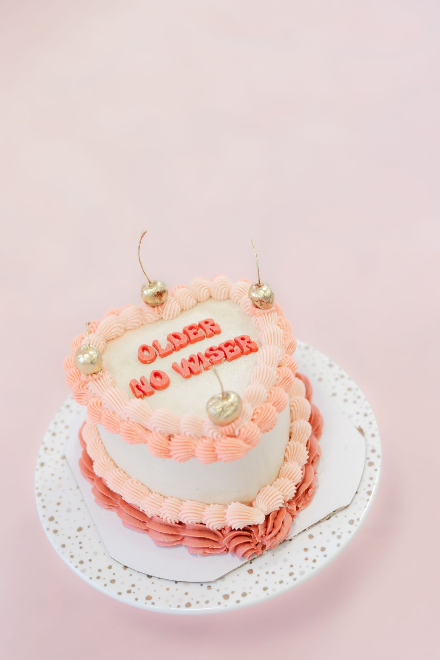 Pink heart-shaped cake with 'older & wiser' text decorated with delicate roses on a lace doily against a pastel background.