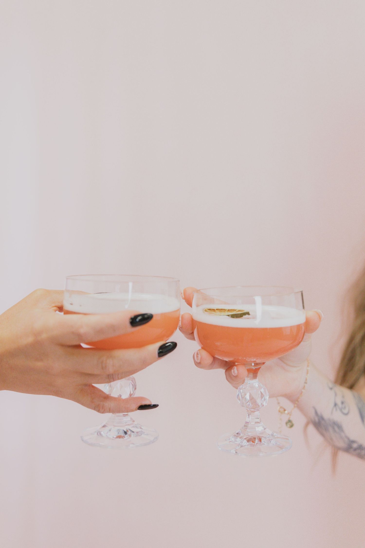 Two hands with black nail polish clink champagne coupe glasses filled with pink cocktails against a pale pink background.