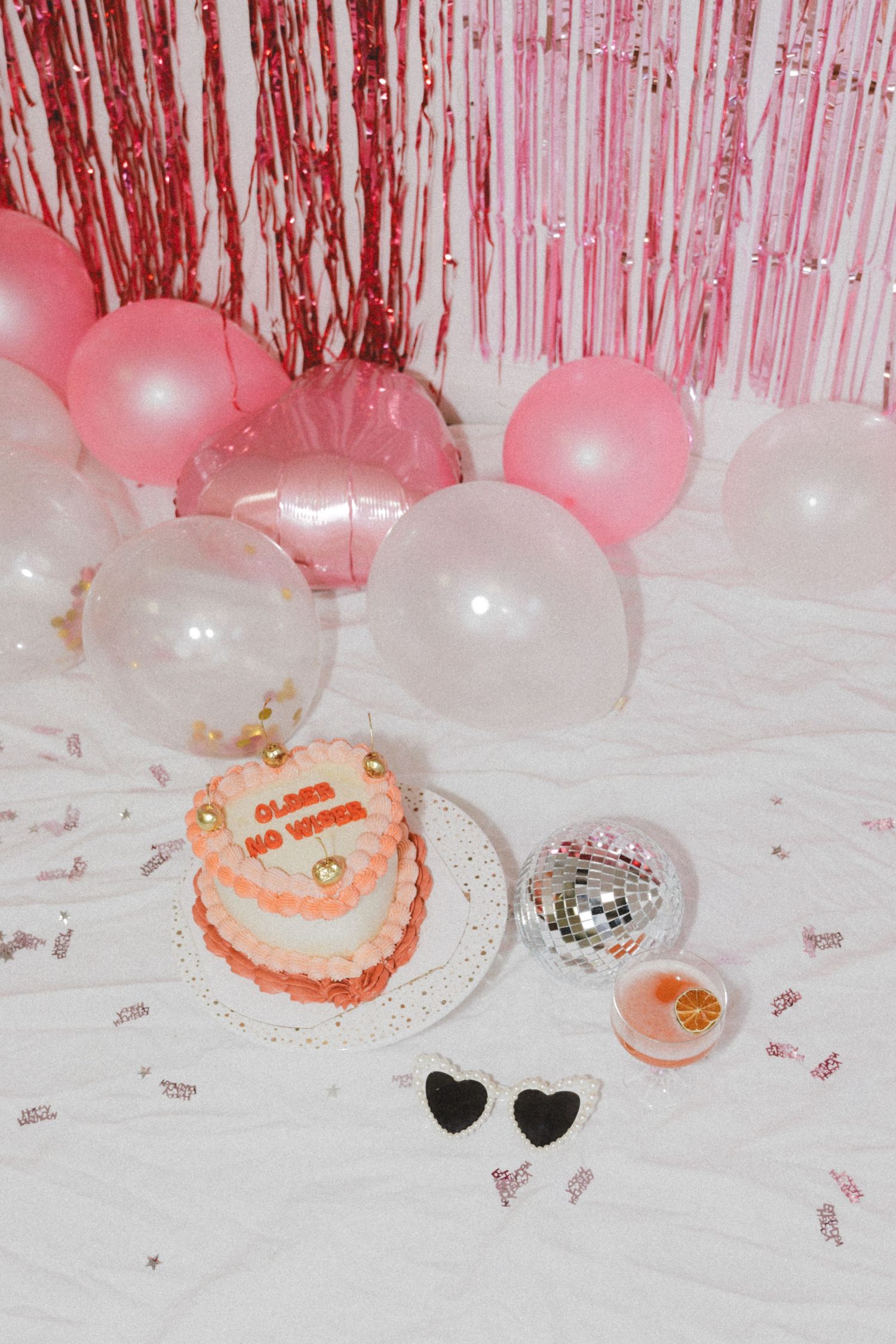 Pink and white balloons with metallic fringe backdrop next to a small cake and disco ball on white surface.