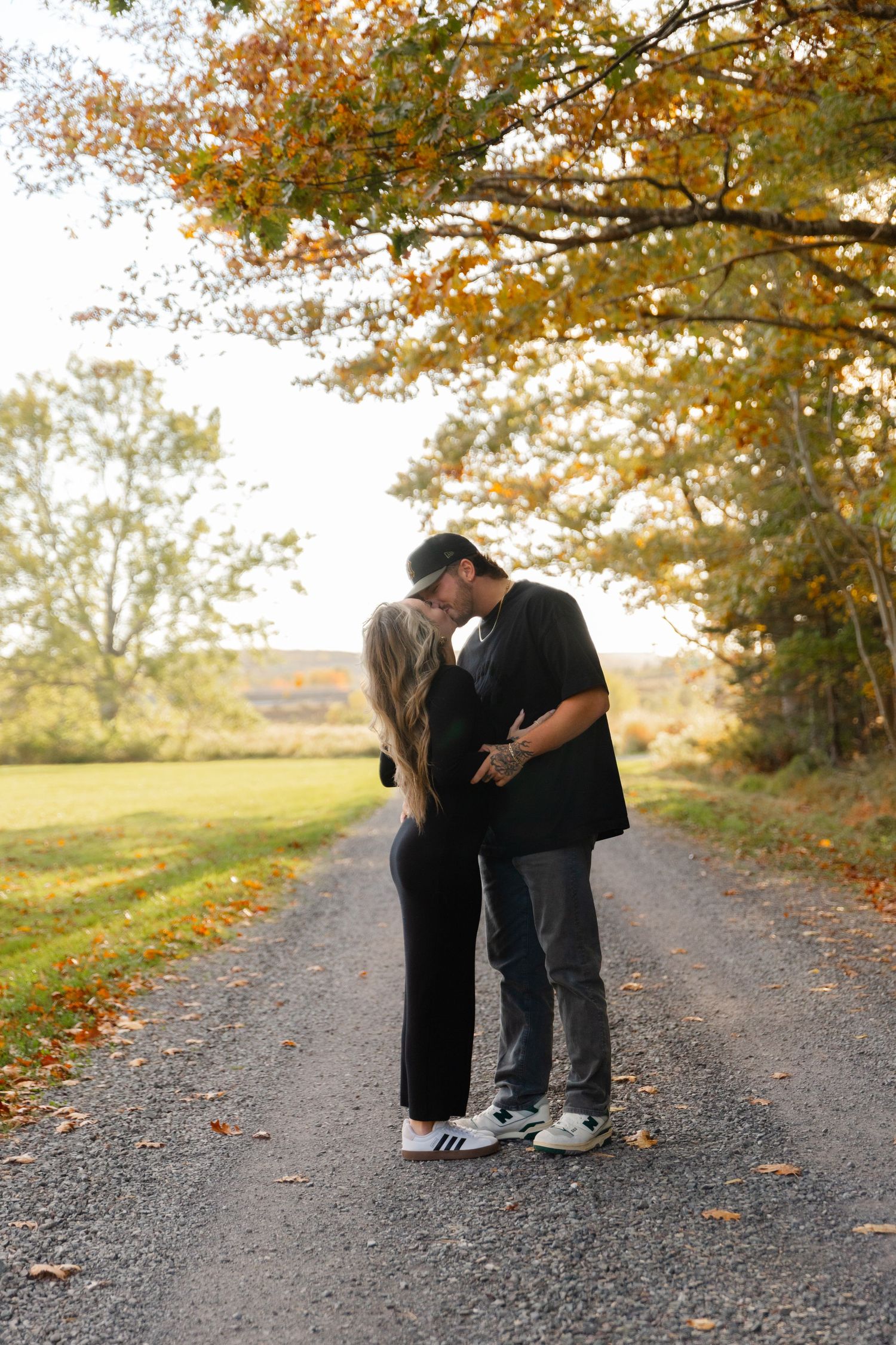 A couple shares a tender moment on an autumn path lined with golden trees as fallen leaves scatter around them.