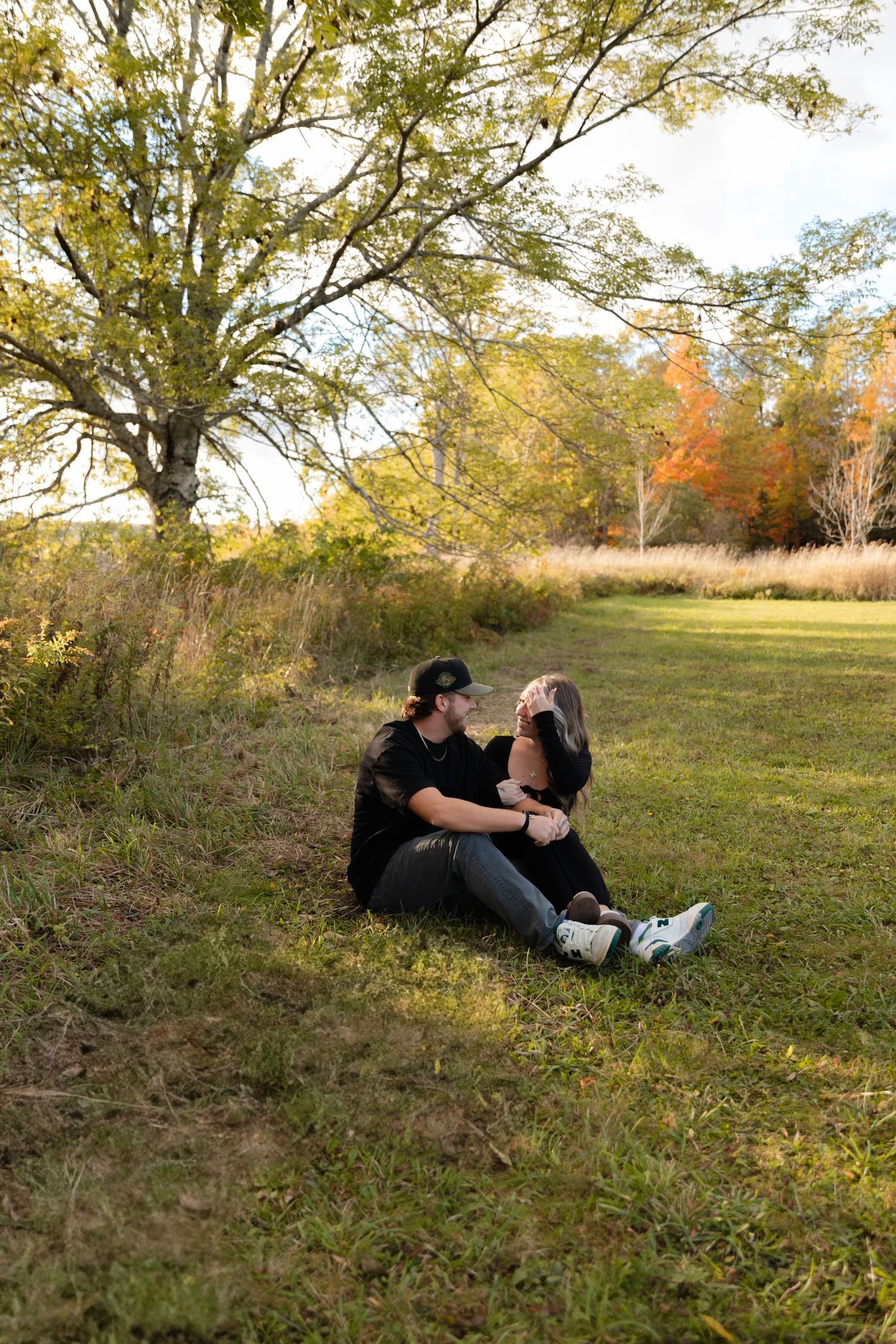 A couple sits together on grass during autumn with fall foliage and trees in the background during golden hour.