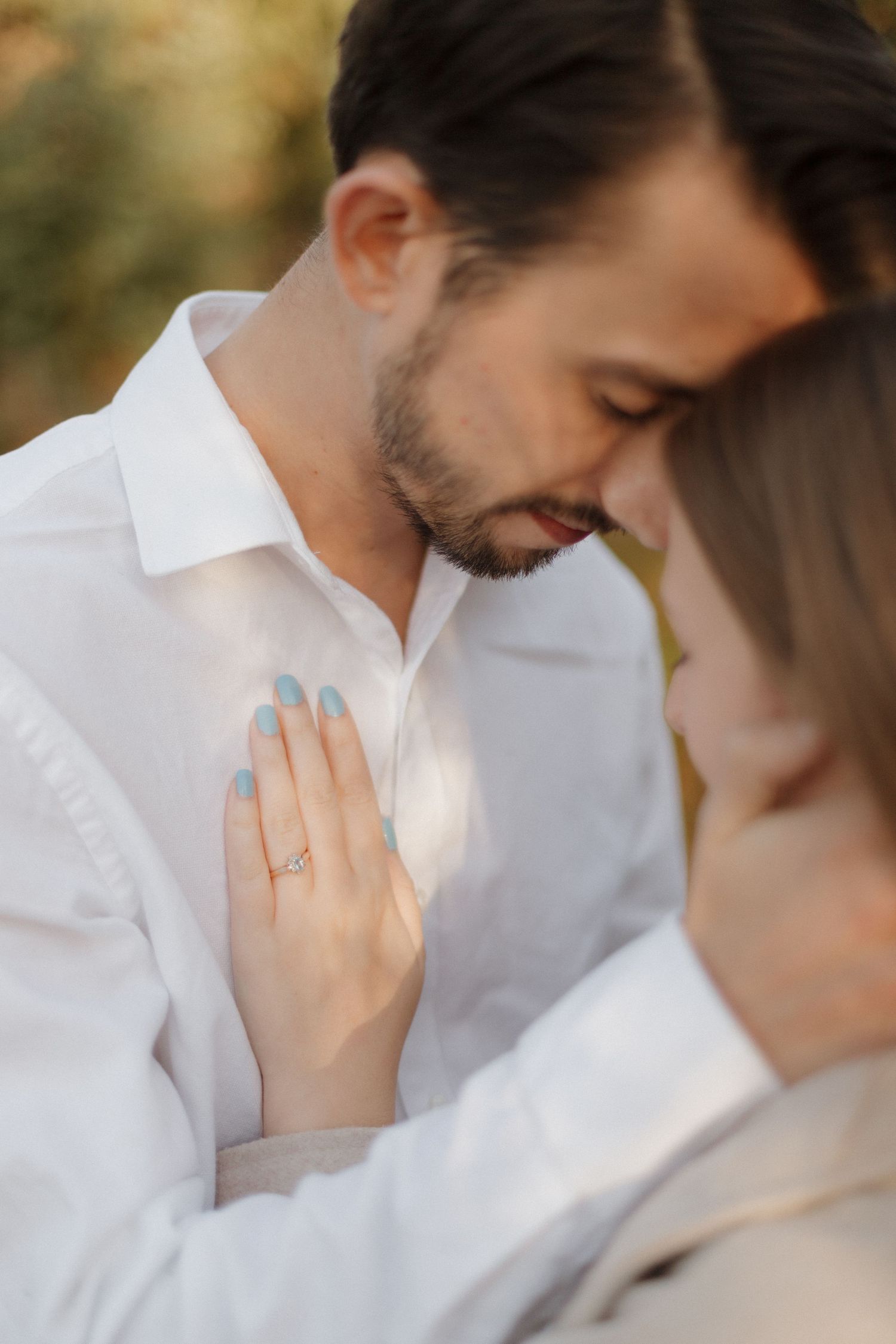 An intimate romantic moment between a couple in white shirts as a hand with an engagement ring rests on his chest.