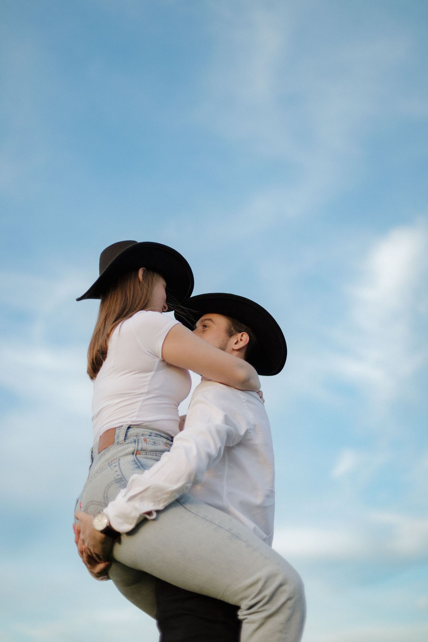A romantic country western couple in cowboy hats embrace against a blue cloudy sky.
