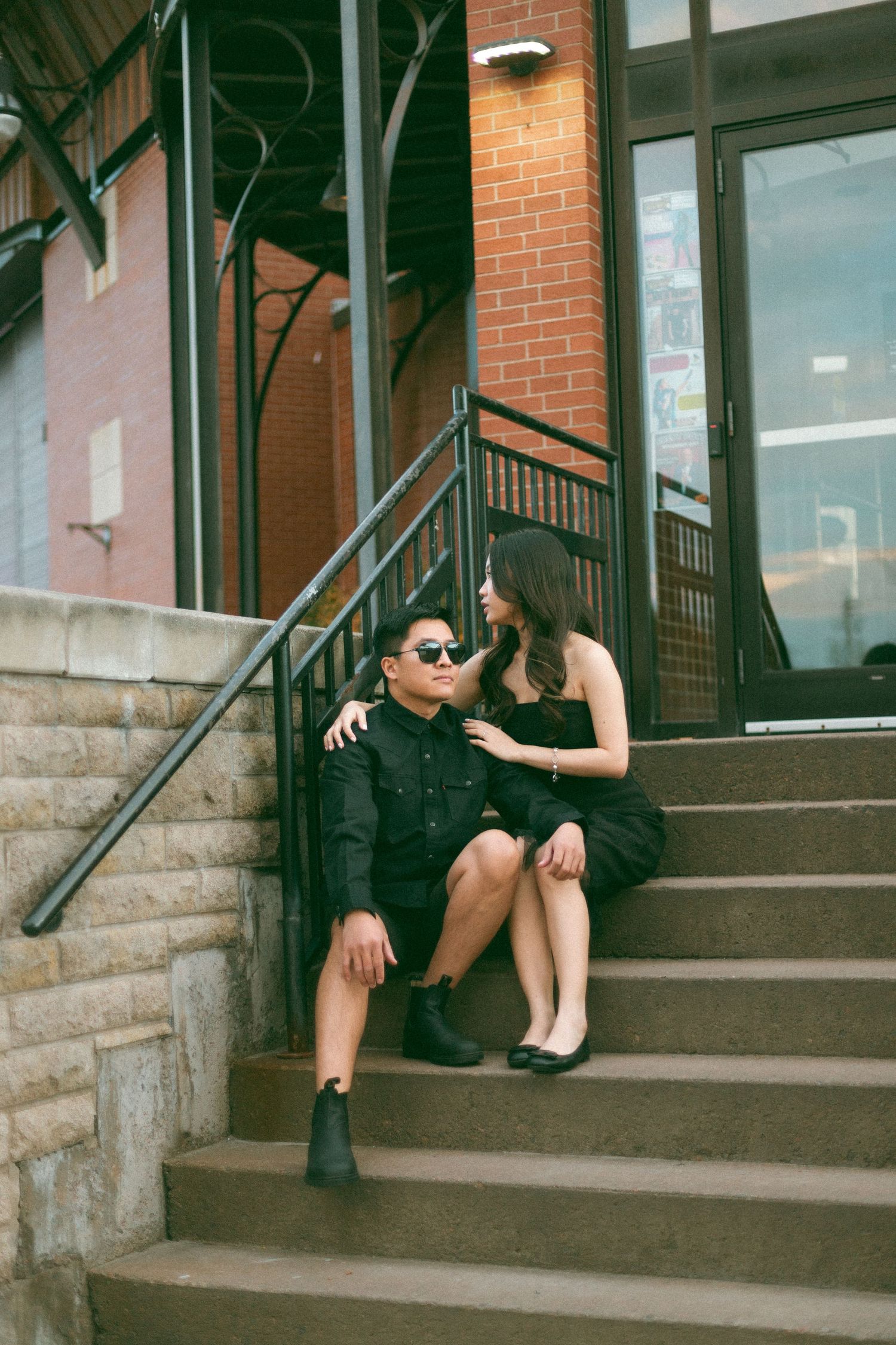 Two people in dark outfits sit casually on concrete steps outside a building with brick and metal details.